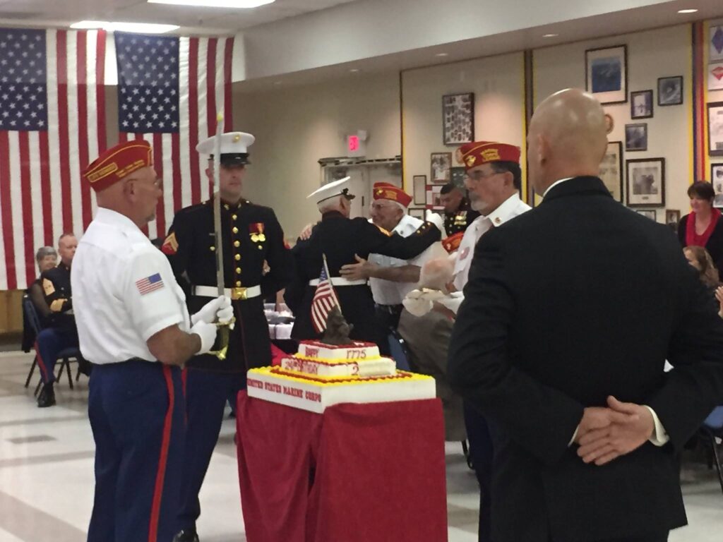 In the background, Commandant Phil Carper presents the first piece of cake to the oldest Marine present, WWII Veteran Do